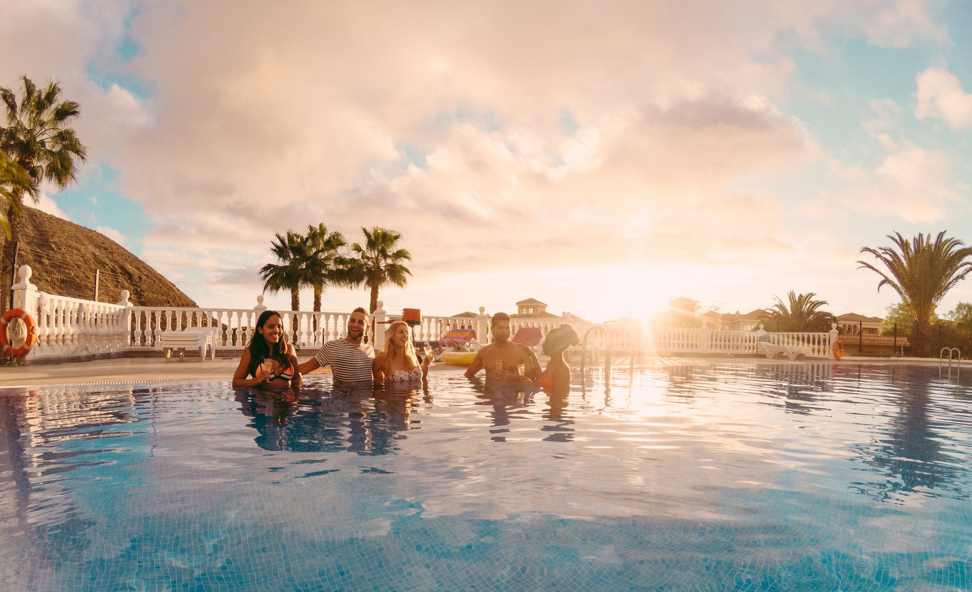 Happy friends drinking champagne in pool party at sunset - Rich people having fun in exclusive tropical vacation - Holiday and friendship concept - Main focus on left guys - Original sun colors tones